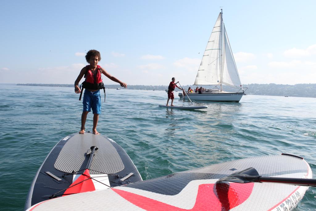 Un enfant joue sur une planche, au loin, un voilier prend le large